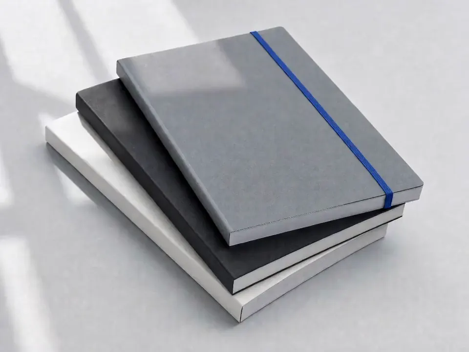 Three softcover workbooks stacked, the top one held by a cobalt elastic band, light passing over the covers.